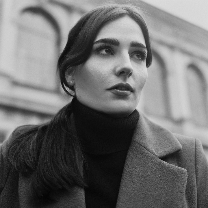 A woman in a coat and turtleneck looks up and to the right, with arched windows in the background.