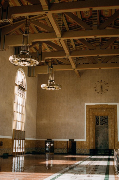 Sunlight streams through a tall arched window onto the tiled floor of a large hall with ornate chandeliers and a patterned ceiling.