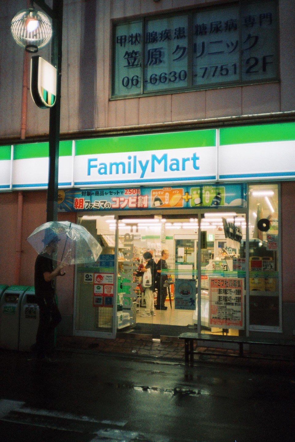 A man with an umbrella stands outside a FamilyMart convenience store at night, with reflections on the wet pavement.