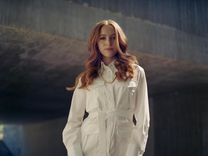 A woman with long, wavy red hair and a white jumpsuit stands in front of a concrete structure.