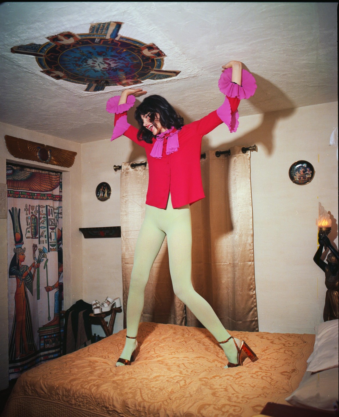 A woman in a pink shirt and green tights stands on a bed in a room with Egyptian-themed decor.