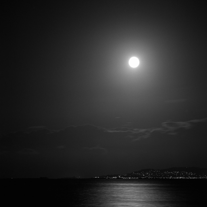 A full moon shines brightly over the sea, with city lights visible on the distant shore.