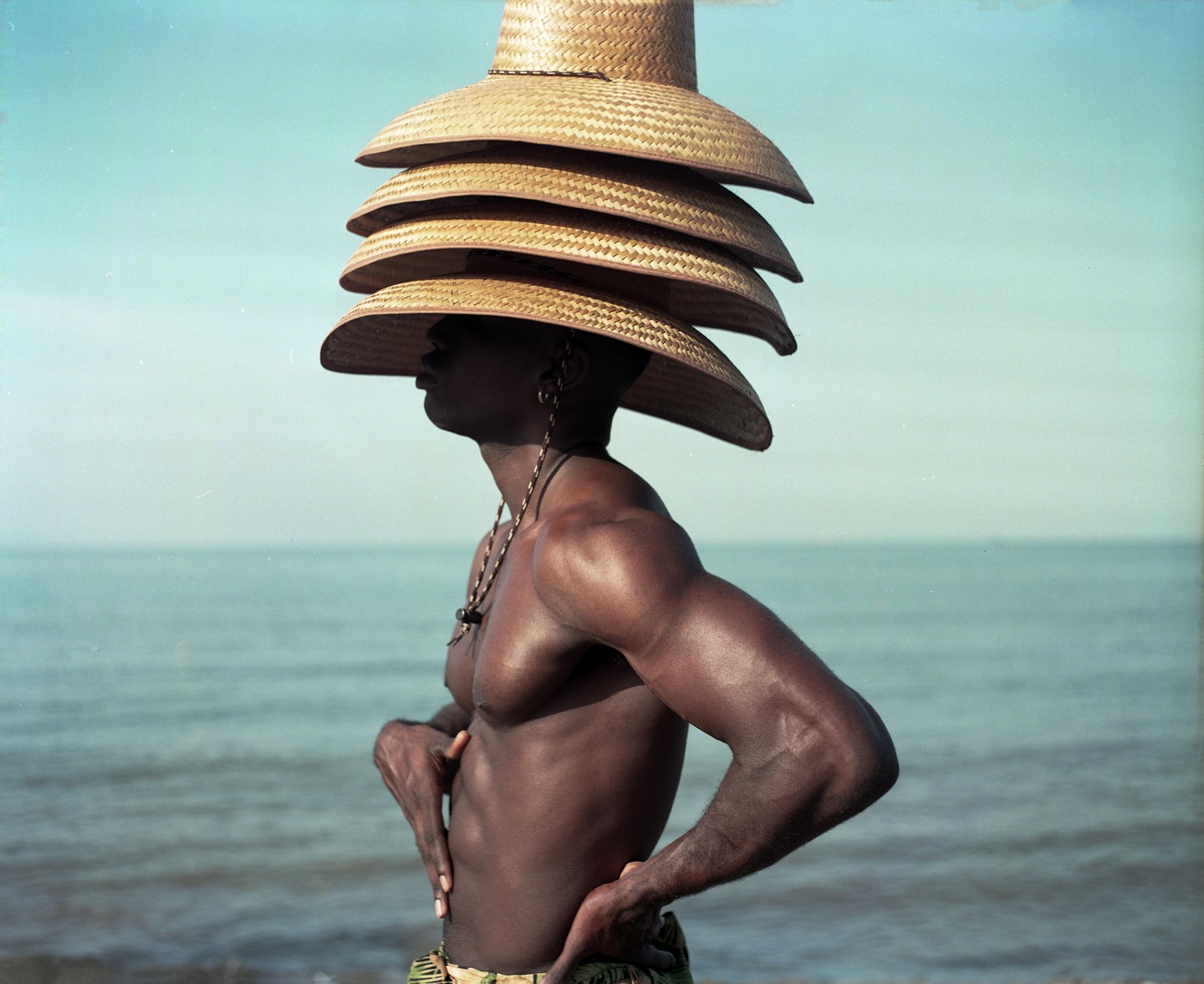 A man with a stack of straw hats on his head stands by the ocean.