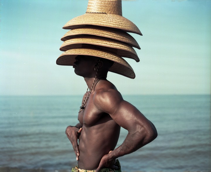 A man with a stack of straw hats on his head stands by the ocean.