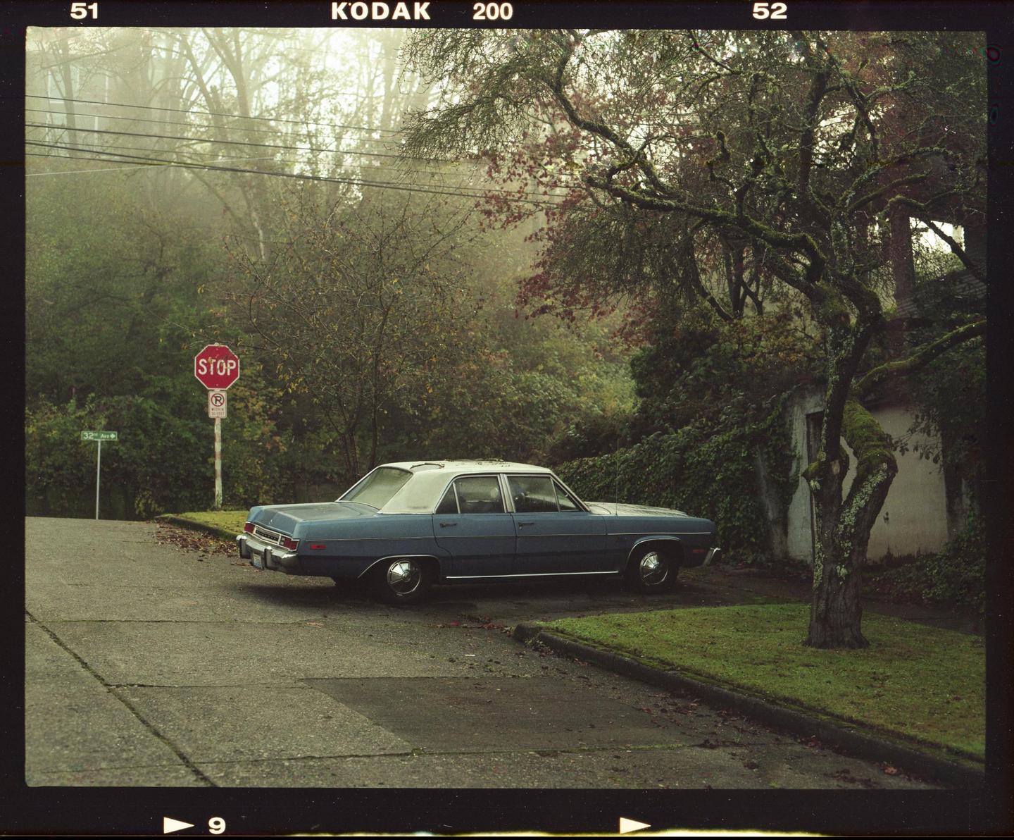 A blue car is parked on a street corner next to a stop sign on a foggy morning.