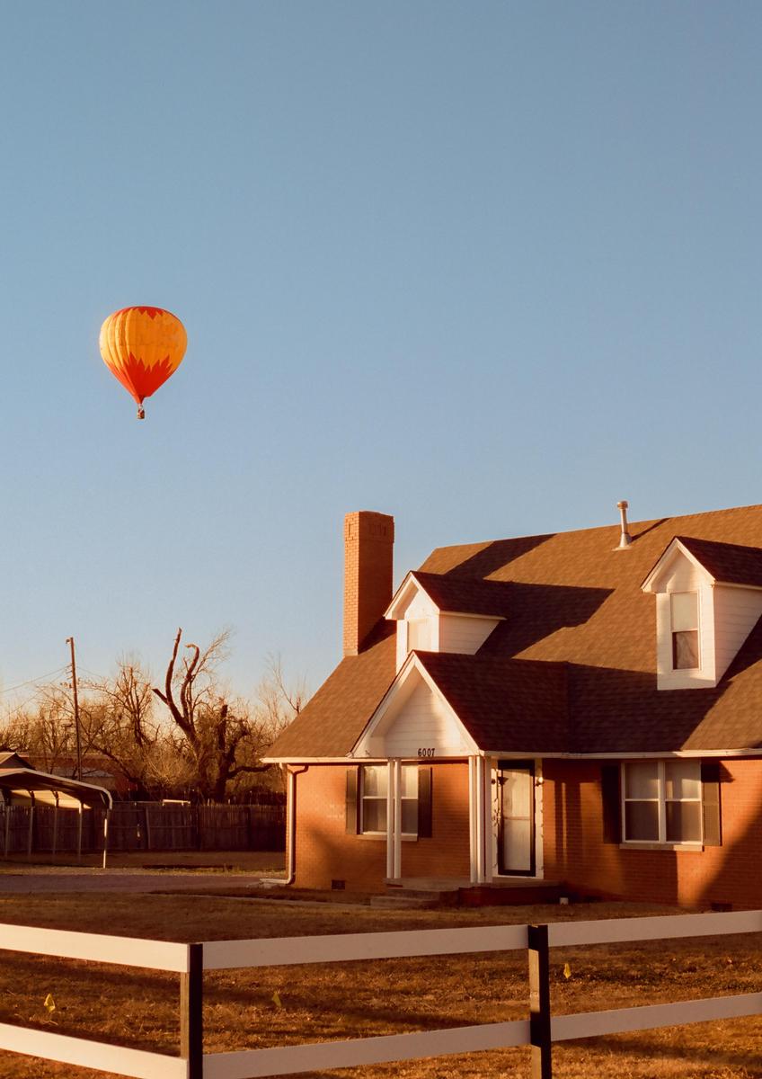 An orange and yellow hot air balloon floats above a brick house with a white fence in the foreground.