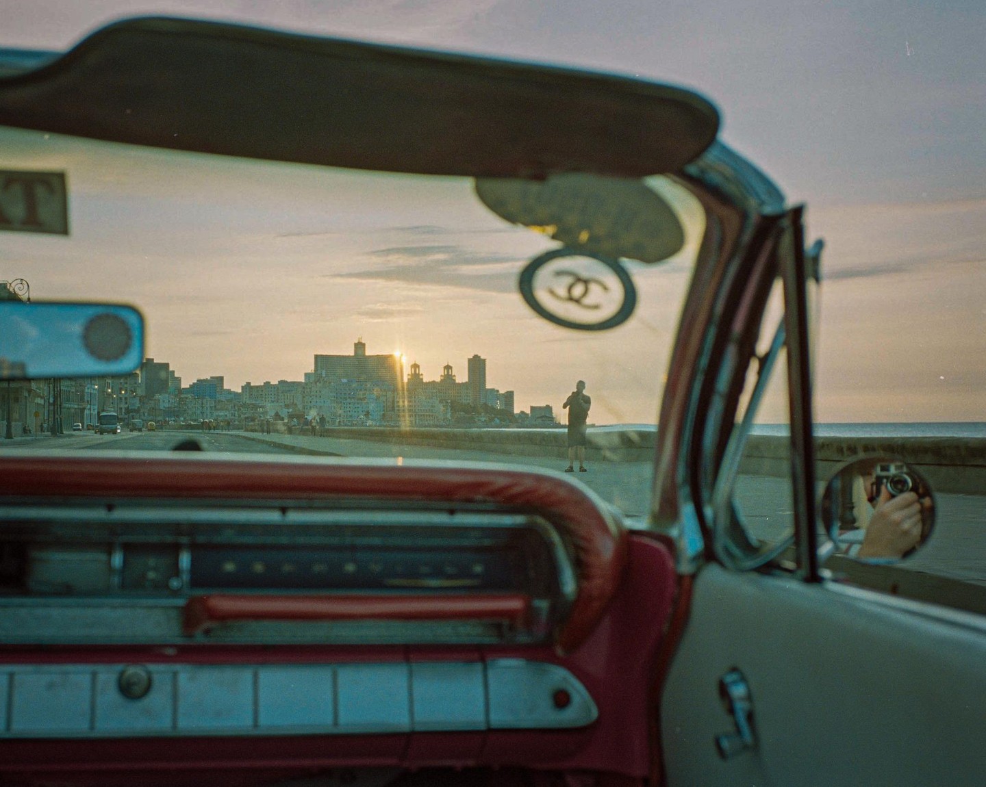 The sun sets over a city skyline as seen from inside a vintage car.