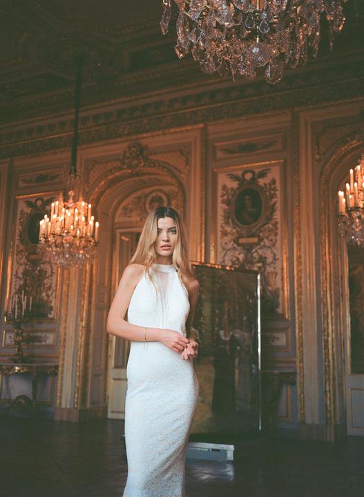 A woman in a white dress stands in a grand room with ornate walls and chandeliers.