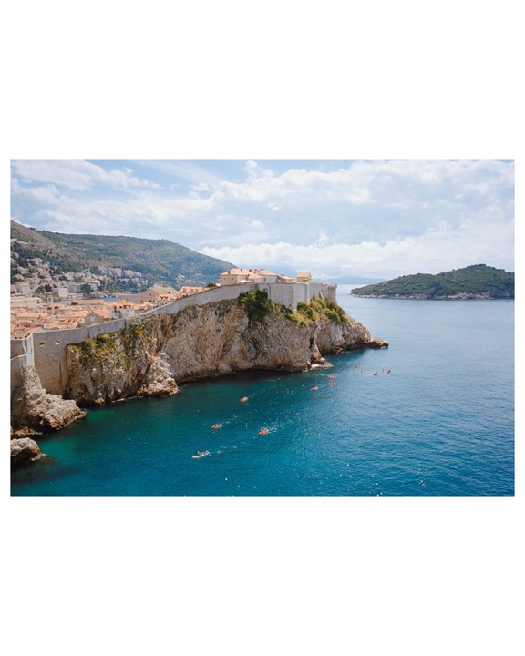 A historic walled city sits atop a cliff overlooking a bay with several small boats and a distant island.