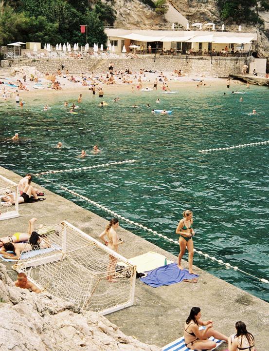 Many people are swimming in the clear blue water and sunbathing on the beach and concrete pier.