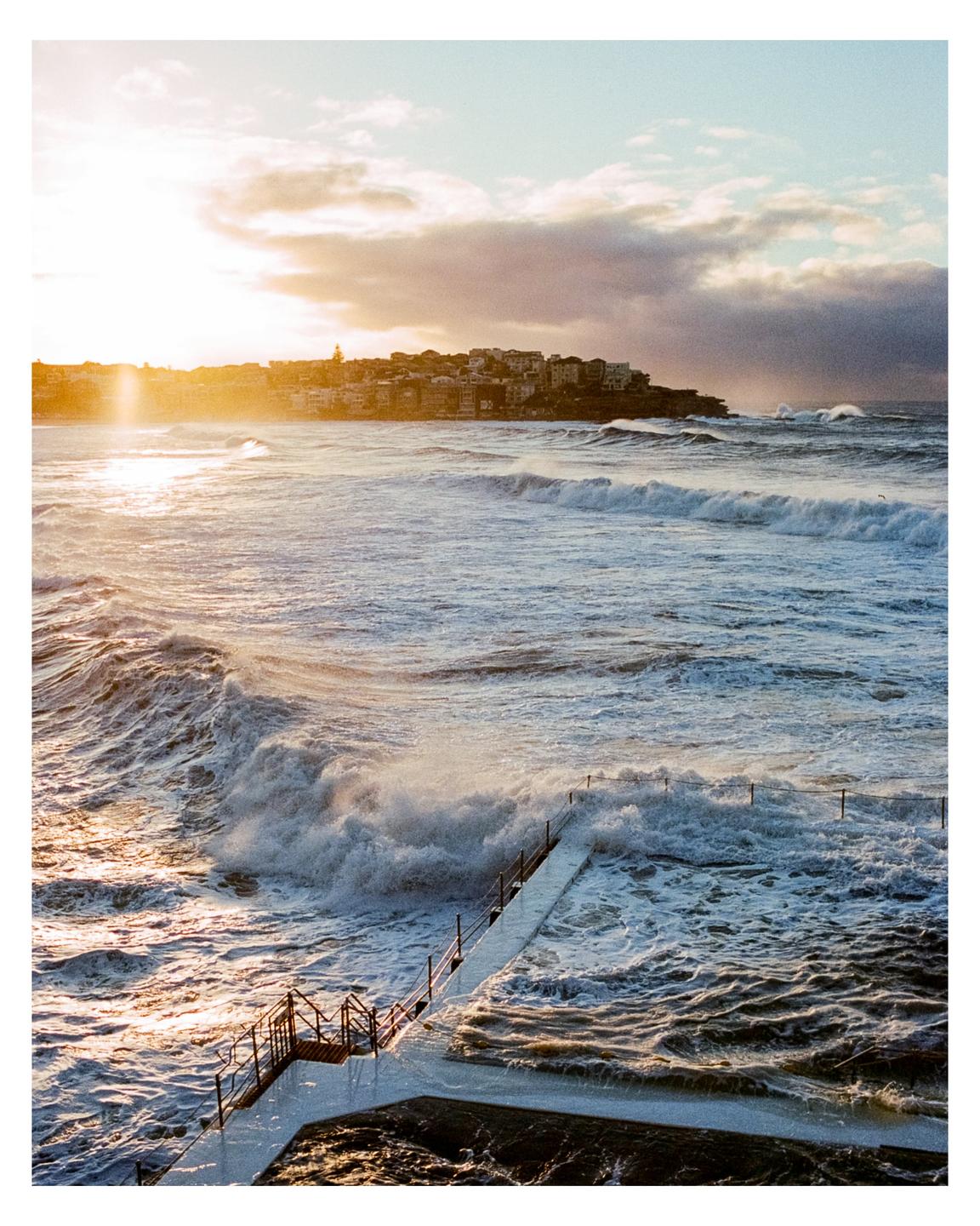 Waves crash into an ocean pool at sunset with a town on a hill in the background.