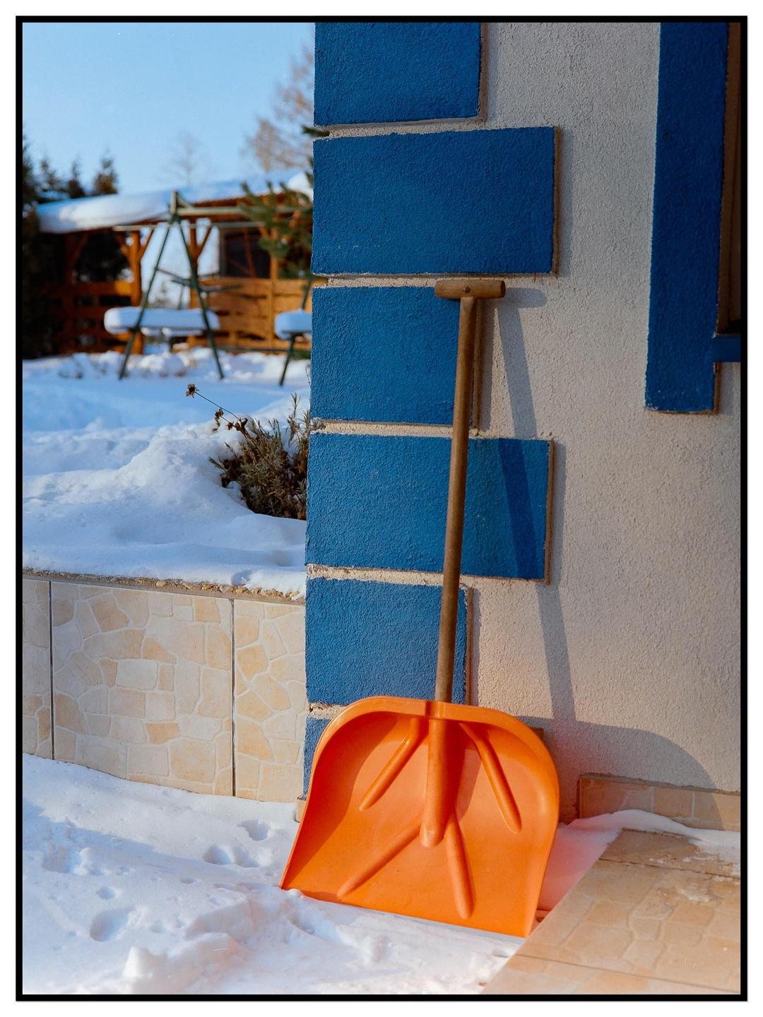 An orange snow shovel leans against a blue and white wall next to a snow-covered ground.