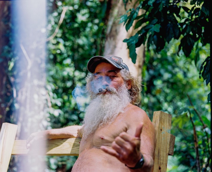 A man with a long white beard and a cap smokes a cigar and makes a shaka sign.