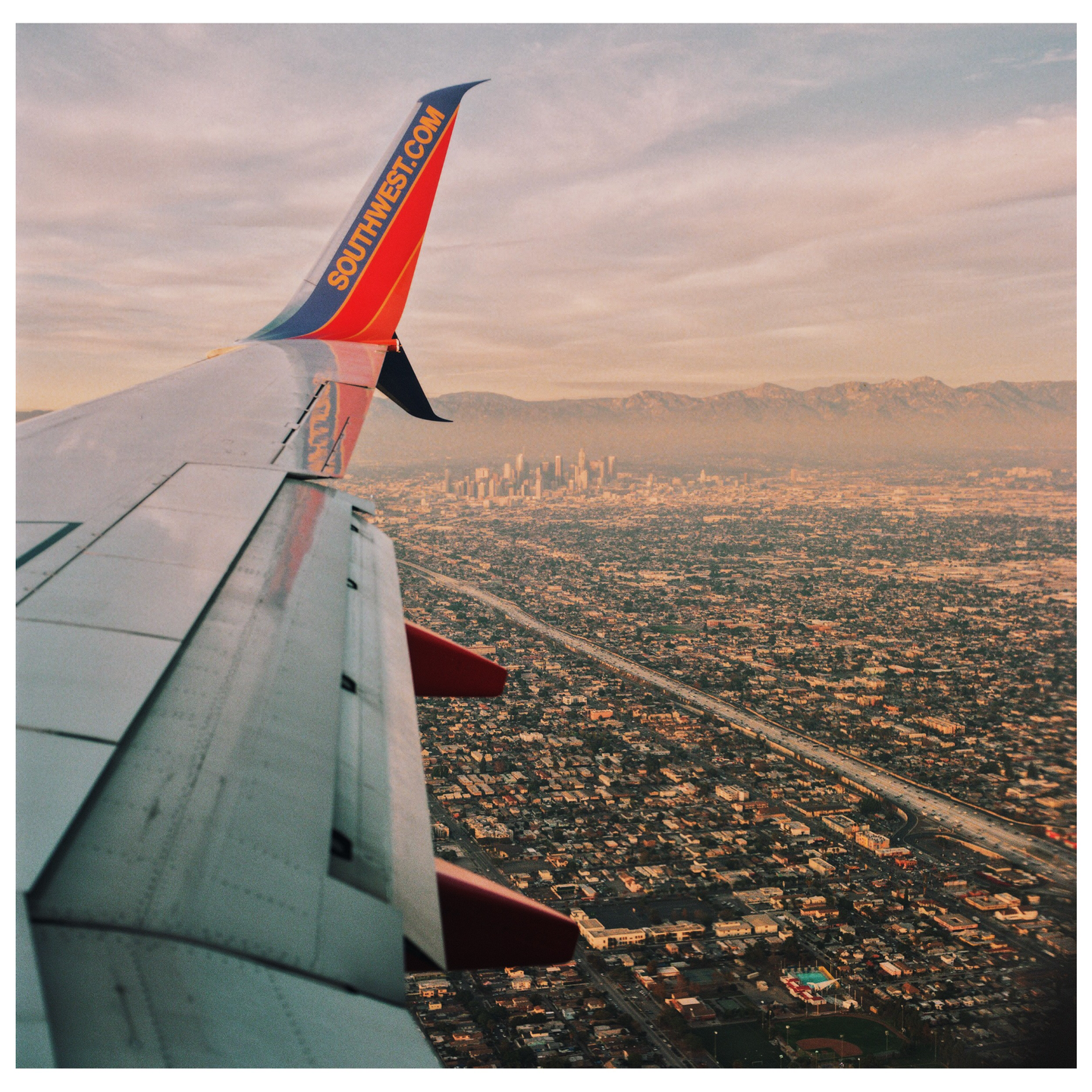 An airplane wing is visible over a sprawling city with mountains in the distance.