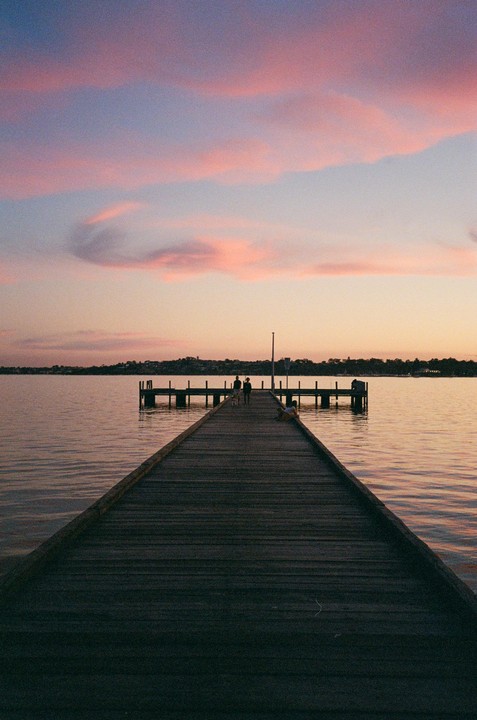 A long wooden pier extends into a calm body of water under a sky with pink clouds at sunset.