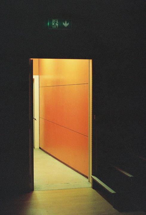 A dark hallway leads to an open doorway revealing a brightly lit orange wall and a green exit sign is visible above the doorway.