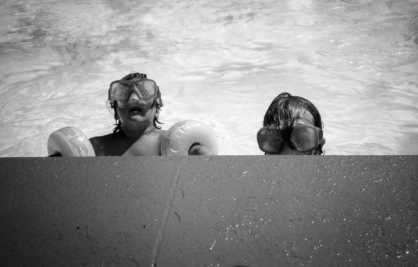 Two children wearing goggles are in a swimming pool, with one child holding an inflatable arm band.