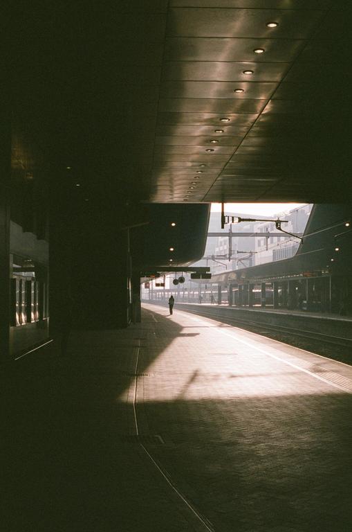 A lone person stands on a sunlit train platform with tracks stretching into the distance.