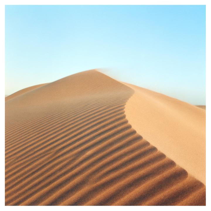 A large sand dune with ripples on its surface rises against a clear sky.