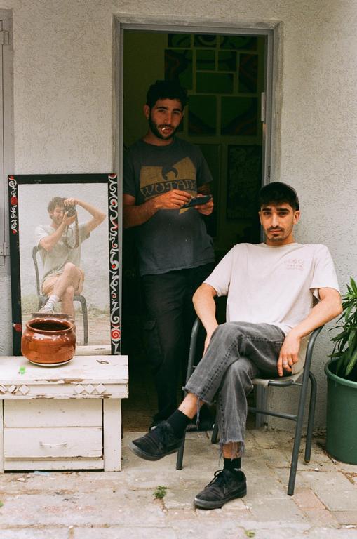 Two men are visible, one standing in a doorway and the other sitting in a chair, with a mirror reflecting a third man holding a camera.