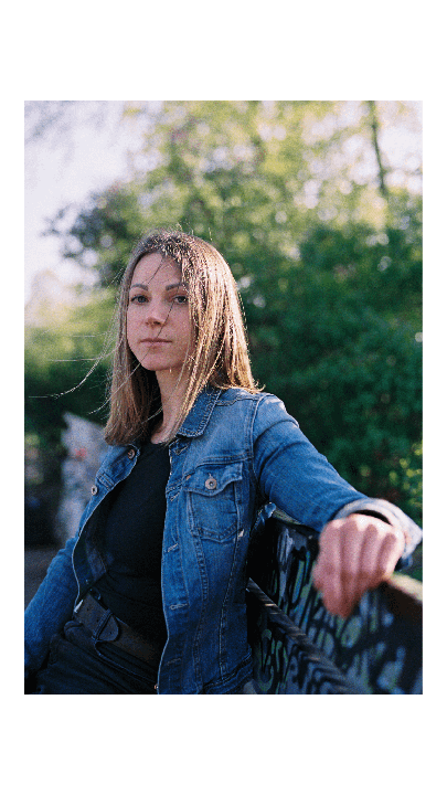 A woman in a denim jacket leans on a railing with graffiti, looking directly at the viewer.