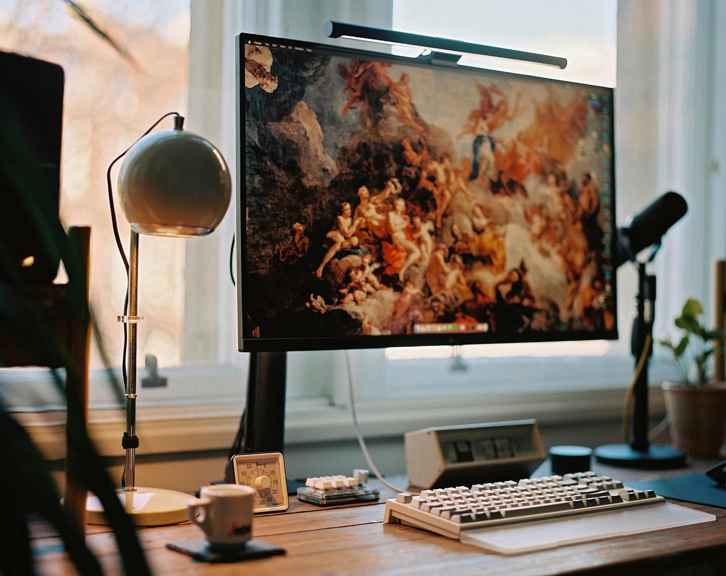 A computer monitor displaying a classical painting is on a wooden desk with a keyboard, a lamp, and a mug.