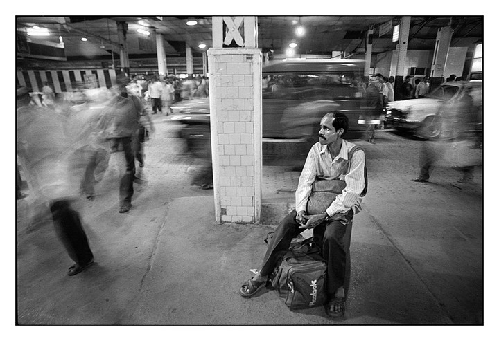 A man sits on a bag next to a pillar while blurred people and vehicles move around him at Howrah Railway Station.
