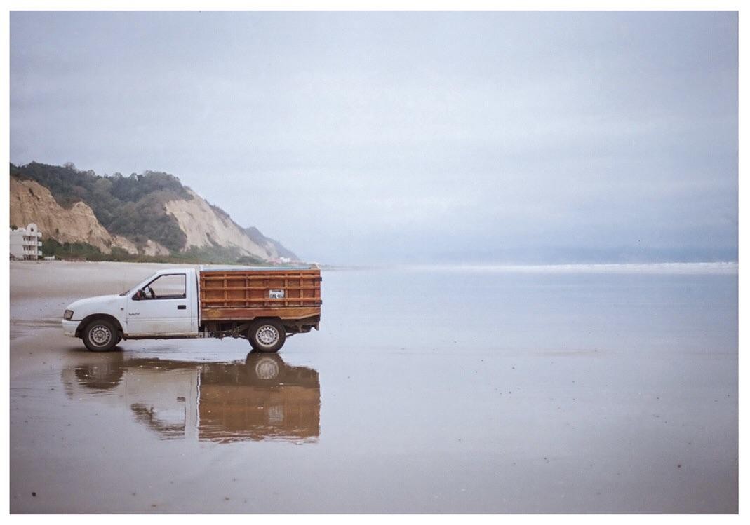 A white pickup truck with a wooden bed is parked on a wet beach with cliffs and buildings in the background.