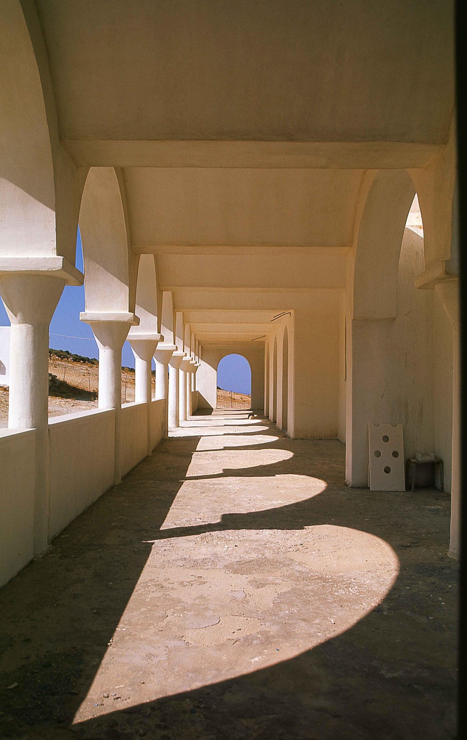 A long, arched hallway with pillars on the left casts shadows on the ground, leading to an archway at the end.