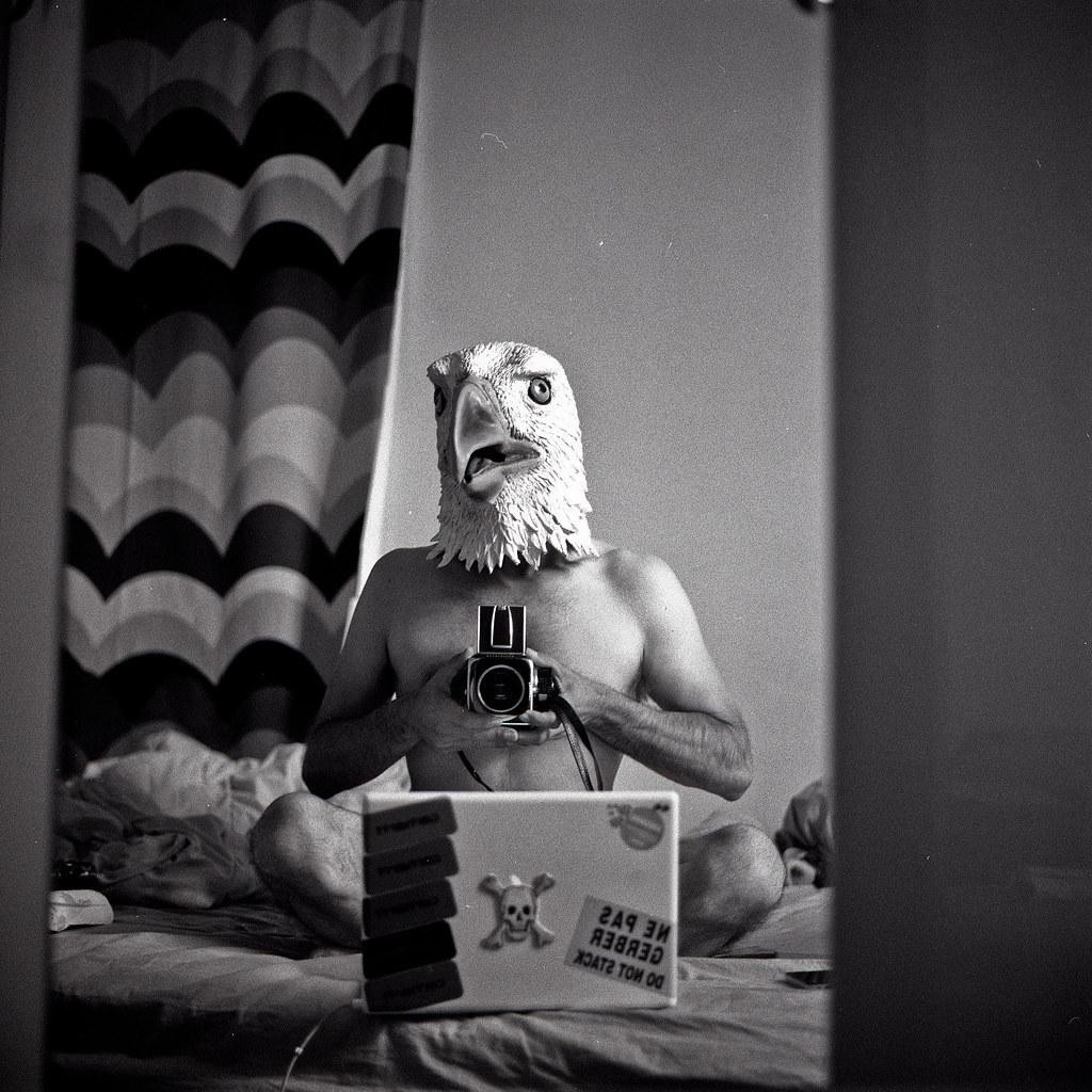 A shirtless man wearing an eagle mask sits on a bed, holding a camera and looking at his reflection in a mirror, with a laptop in front of him.