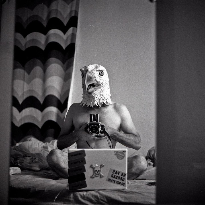 A shirtless man wearing an eagle mask sits on a bed, holding a camera and looking at his reflection in a mirror, with a laptop in front of him.