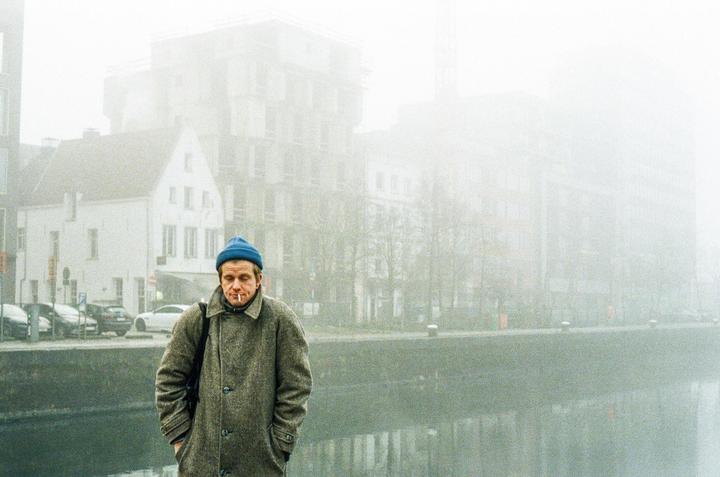 A man wearing a blue beanie and a coat stands by a canal on a foggy day.