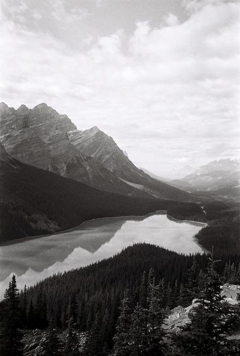A black and white landscape shows a lake surrounded by mountains and forest under a cloudy sky.