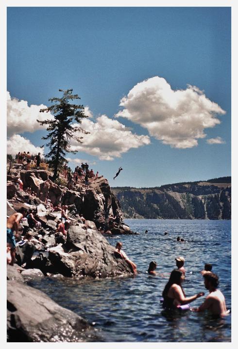 A person jumps from a rocky cliff into a lake where other people are swimming, with mountains in the background under a blue sky with clouds.