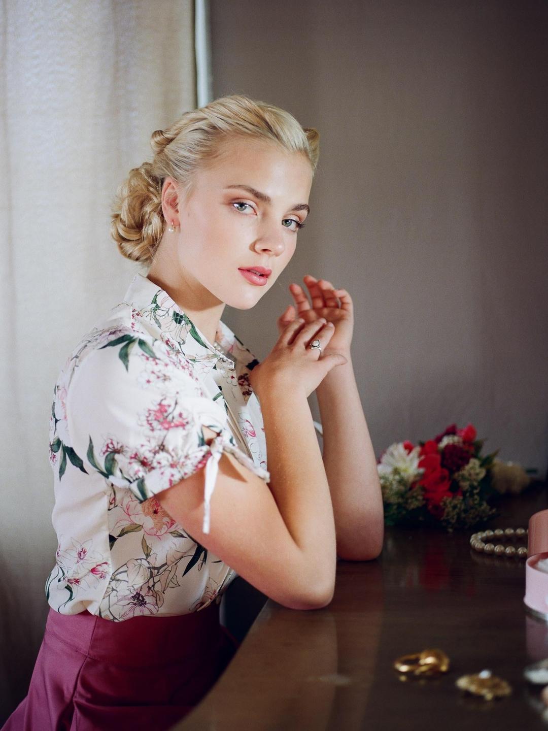 A woman with braided hair and a floral shirt sits at a wooden table with her hands clasped.