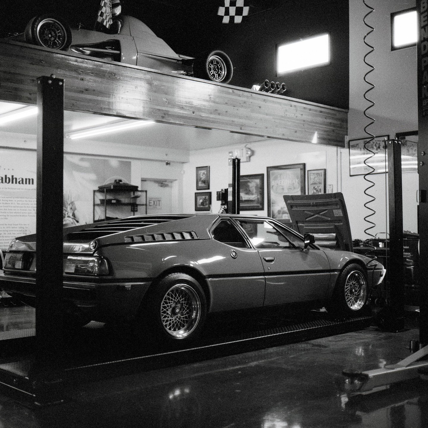 A sports car with its hood open sits on a lift in a garage, with another race car visible on a shelf above.