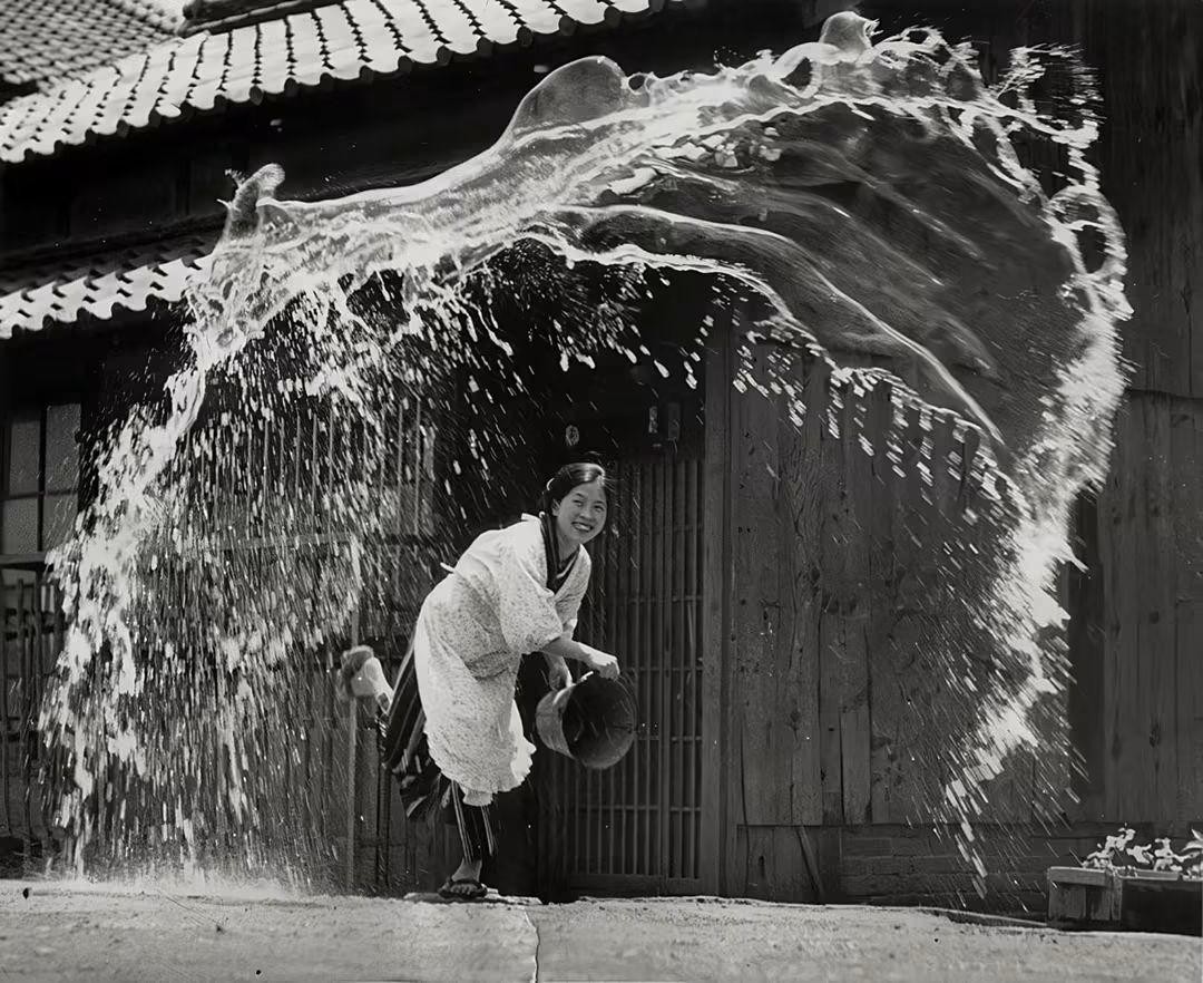 A woman in traditional clothing smiles while being splashed by a large volume of water in front of a wooden building.