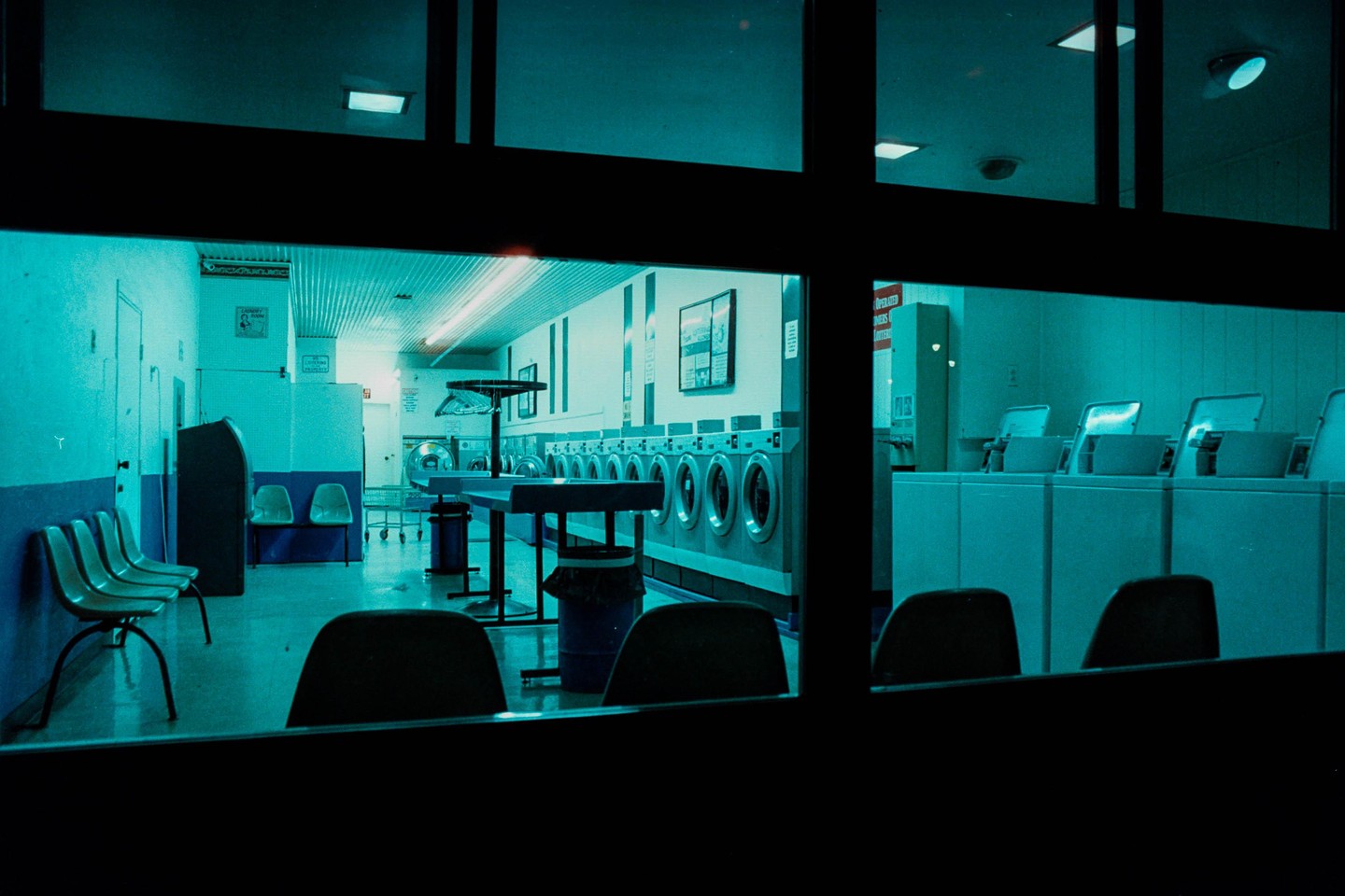 A laundromat interior is viewed through a window at night, illuminated by a strong blue light.