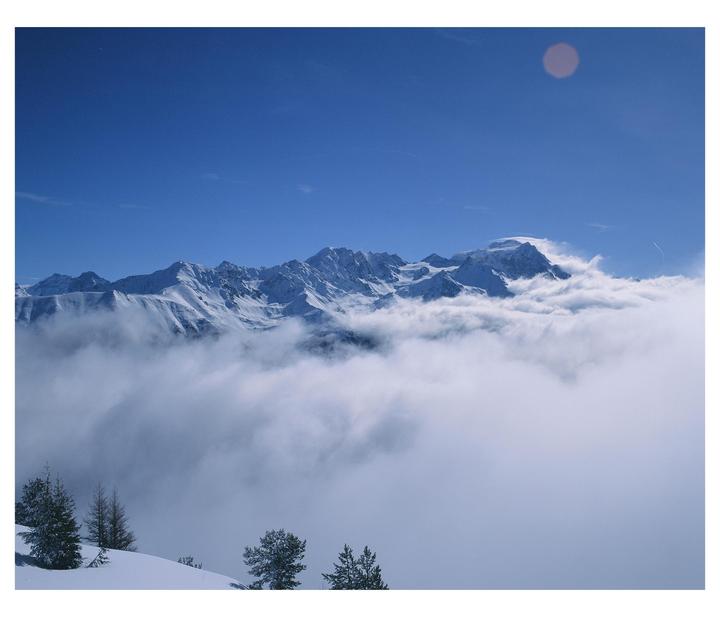 Snow-covered mountains rise above a sea of clouds under a clear blue sky.