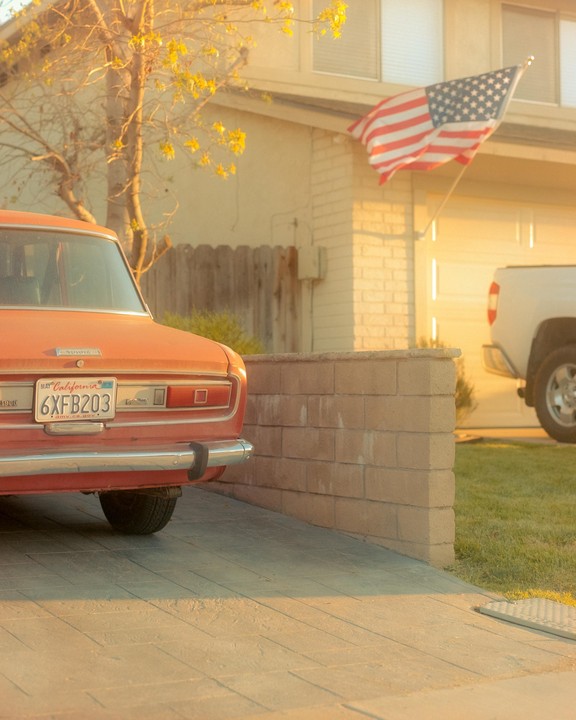 An orange car is parked in a driveway with an American flag hanging from a house in the background.