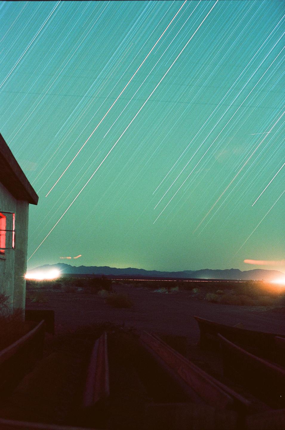 A building with a red window is on the left, with star trails in the sky and light trails on the horizon.