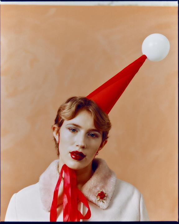 A woman with red lipstick and a red party hat with a white ball on top looks directly at the viewer.