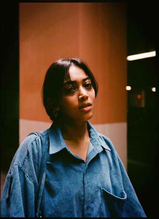 A woman with short dark hair and a blue corduroy shirt looks slightly to the right of the camera.