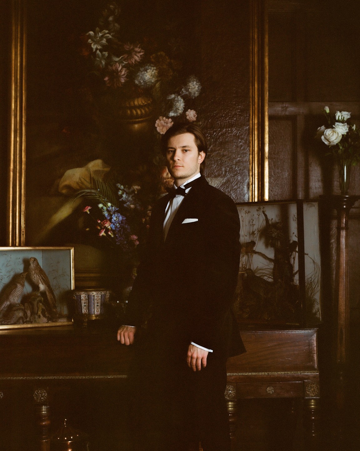 A man in a tuxedo stands in a room with several paintings and a vase of flowers.