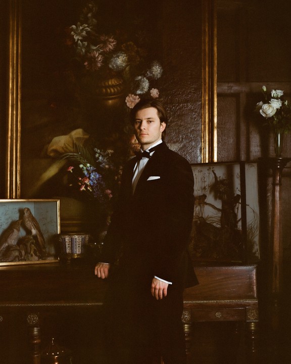 A man in a tuxedo stands in a room with several paintings and a vase of flowers.