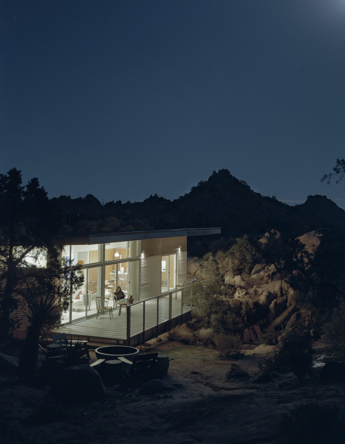 A modern house with a lit interior and a deck sits among rocky hills and desert plants under a night sky.