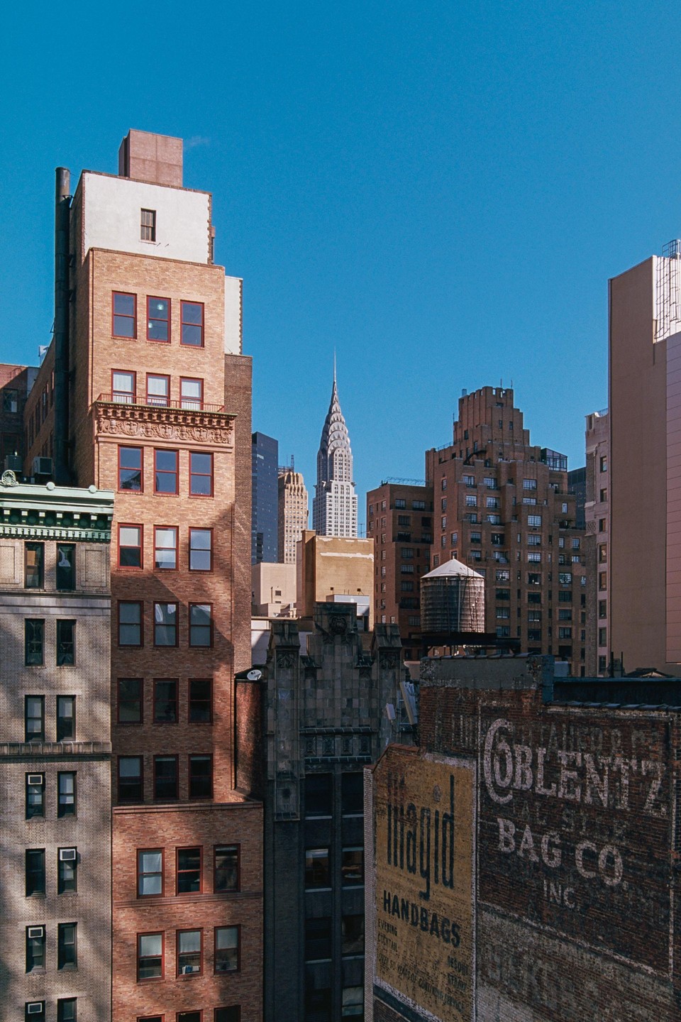 The Chrysler Building rises above other buildings in Midtown, with a water tower and painted signs on brick buildings in the foreground.