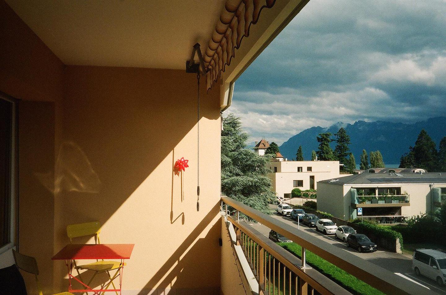 A balcony with a red table and yellow chairs overlooks a street with cars and buildings, with mountains and a lake in the distance under a cloudy sky.