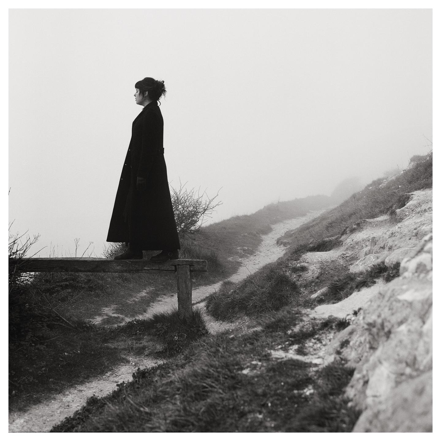 A woman in a long coat stands on a wooden barrier looking out into a foggy landscape with a path winding up a hill.