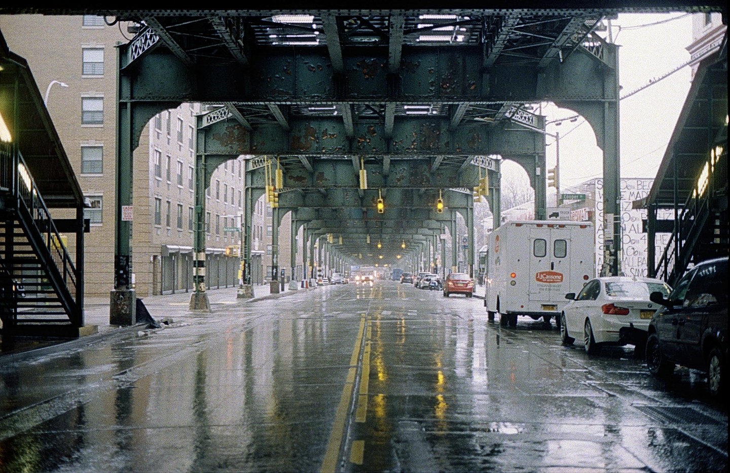 A wet street with cars is visible under a subway overpass in Brooklyn, New York.